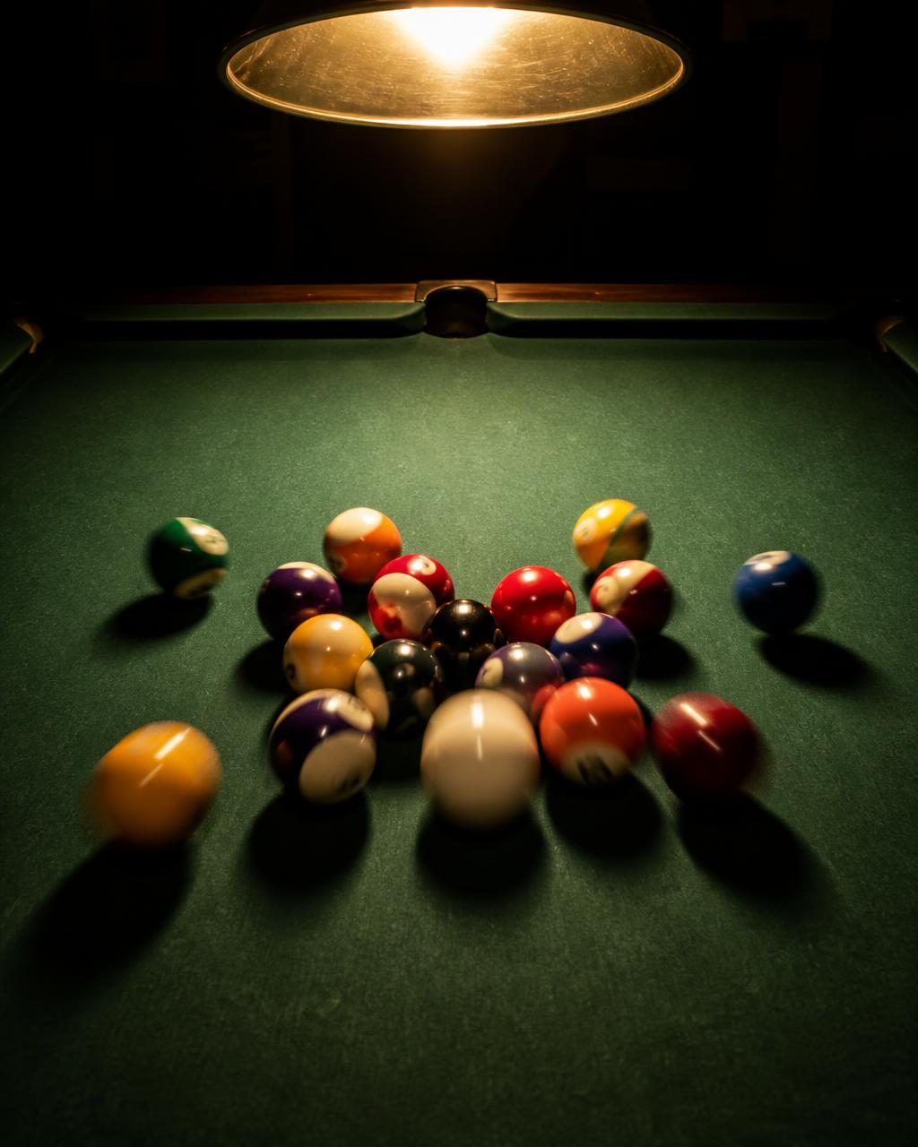An eight ball rack breaking apart under a warm pool table lamp